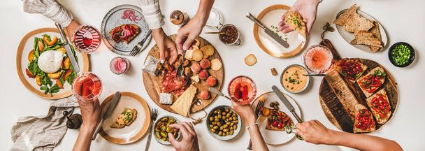 Friends wine and snacks party. Flay-lay of people eating and drinking rose wine over table with cheese, fruit, smoked meat, tomato brushettas, buratta salad, top view. Wine tasting, gathering concept - 写真・画像