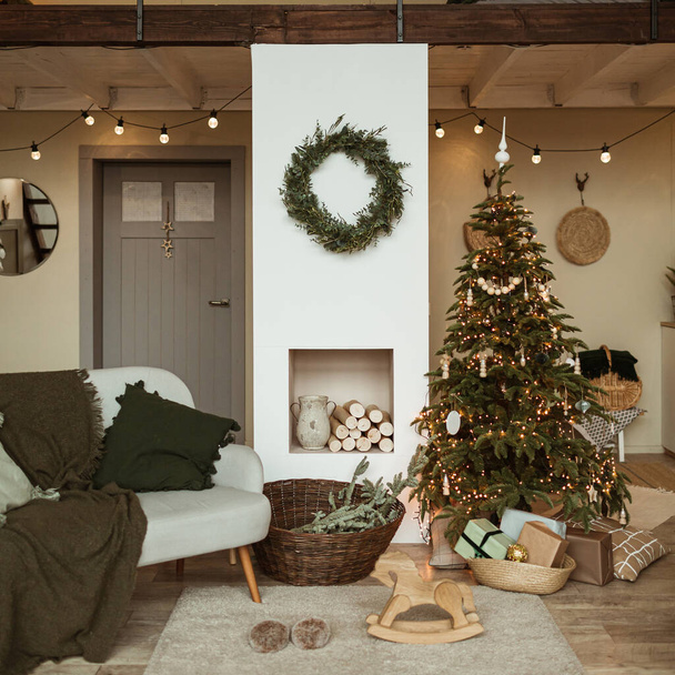 Modern interior design concept. Comfortable cozy living room decorated with Christmas tree with gifts, wreath frame, fireplace, sofa, carpet. Christmas / New Year celebration decorations. - Photo, Image