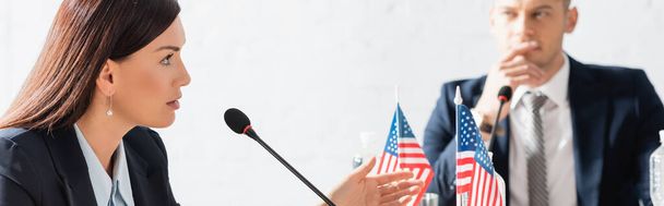 Mujer política haciendo gestos, mientras habla en micrófono, sentada cerca de banderas americanas con un hombre borroso sobre un fondo borroso, pancarta - Foto, Imagen