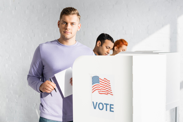 hombre mirando a la cámara mientras sostiene boleta electoral cerca de cabina de votación con bandera americana y letras de voto en un fondo borroso - Foto, Imagen