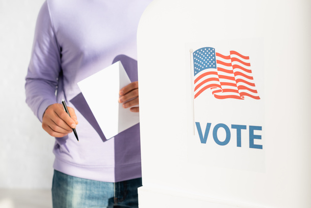 vue recadrée de l'homme tenant un bulletin de vote et un stylo près de l'isoloir avec drapeau américain et inscription du vote - Photo, image