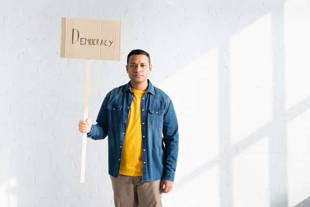 serious indian man holding placard with democracy inscription against white brick wall - Foto, Imagem