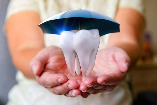 Ένα δόντι κάτω από μια ομπρέλα πάνω από τα χέρια μιας γυναίκας. Προστασία της υγείας, ασφάλιση έννοια, οδοντιατρική, ιατρική περίθαλψη.  - Φωτογραφία, εικόνα