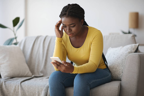 Worried black lady looking at smartphone screen, sitting on couch at home - Foto, Imagen