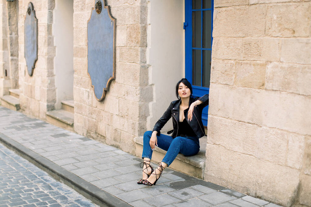 Outdoor portrait of young beautiful fashionable Asian lady, wearing heels, jeans and leather jacket, sitting on stairs of old building with blue door, on a street of the old city. - 写真・画像