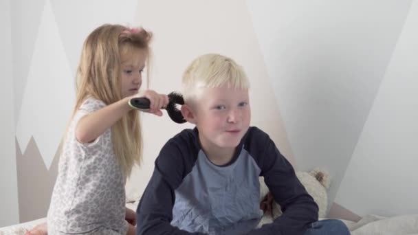 Zabawna Mała Siostra grzebie blond włosy starszemu bratu - Materiał filmowy, wideo