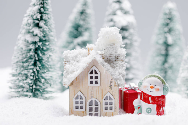 Small decorative snowman with gift near wooden house in fir forest under falling snow - Foto, Imagem
