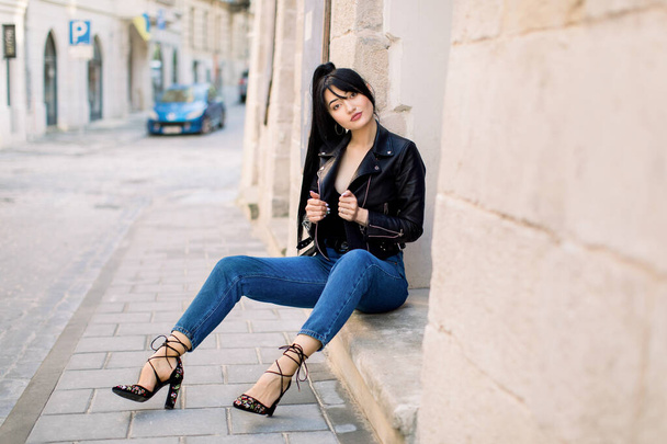 City life, people and fashion concept. Sexy young attractive Asian woman, wearing jeans, heels and black leather jacket, posing to camera outdoors in the sity, sitting on the stairs of building - Fotografie, Obrázek