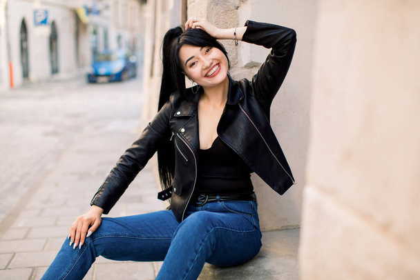 Beautiful sexy young Asian woman with ponytail hair, wearing trendy casual clothes, leather jacket and denim jeans, sitting outdoors on stairs in the city, smiling to camera - Photo, image