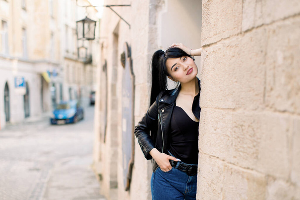 Front view of young charming brunette Asian woman in trendy clothes, posing at the street city against the background of the wall of old building - Foto, Imagen