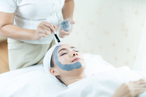 Gezichtsbad. Gezicht huidverzorging. Een Aziatische vrouw maskeert haar gezicht in een spa salon. Hand die een borstel vasthoudt. - Foto, afbeelding