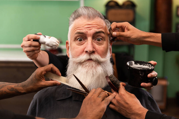 volwassen man met witte baard kijken naar camera in kapperszaak met kapper handen met knippen en scheren instrumenten, borstel, schaar, kam, scheermes. hipster stijl - Foto, afbeelding