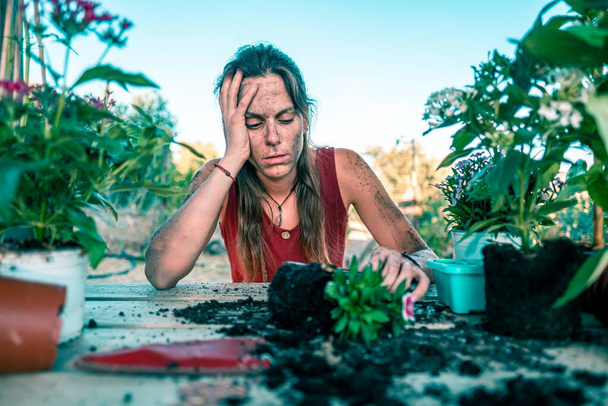 Young caucasian woman wearing a red dress surrounded by plants and pots with a pruning shears on a wooden table - Photo, Image