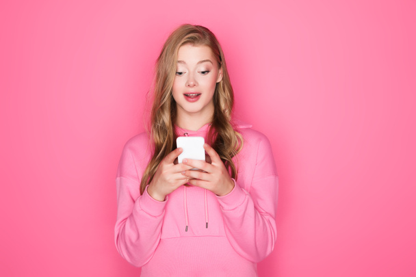 excitada hermosa mujer con la boca abierta celebración de teléfono inteligente sobre fondo rosa - Foto, Imagen