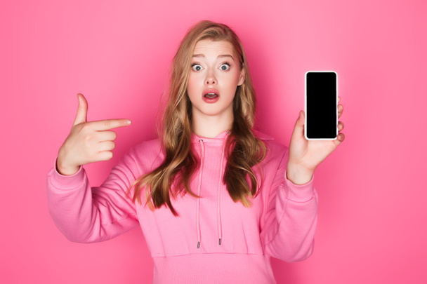 impactado hermosa mujer apuntando al teléfono inteligente sobre fondo rosa - Foto, imagen