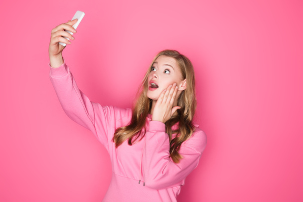 shocked beautiful woman taking selfie on smartphone on pink background - Fotoğraf, Görsel