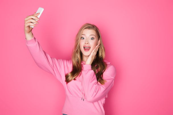 shocked beautiful woman taking selfie on smartphone on pink background - Zdjęcie, obraz