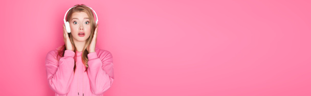 σοκαρισμένη όμορφη γυναίκα ακούγοντας μουσική σε ακουστικά σε ροζ φόντο, πανό - Φωτογραφία, εικόνα