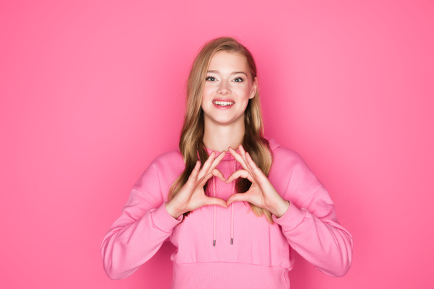 beautiful young woman in hoodie showing heart with hands on pink background - Фото, изображение