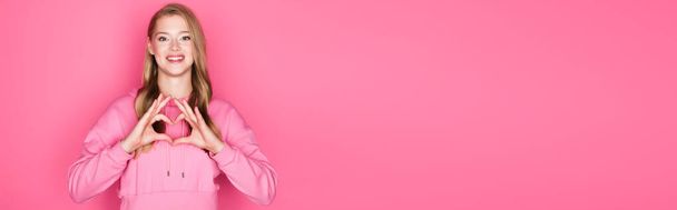beautiful young woman in hoodie showing heart with hands on pink background, banner - Fotoğraf, Görsel