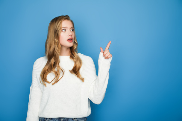 shocked blonde beautiful woman in sweater pointing with finger isolated on blue - Fotografie, Obrázek