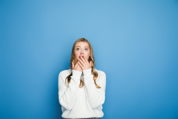 σοκαρισμένη ξανθιά όμορφη γυναίκα σε πουλόβερ που καλύπτει το στόμα απομονώνονται σε μπλε - Φωτογραφία, εικόνα