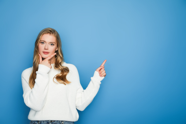 pensive blonde beautiful woman in sweater pointing with finger isolated on blue - Foto, Imagen