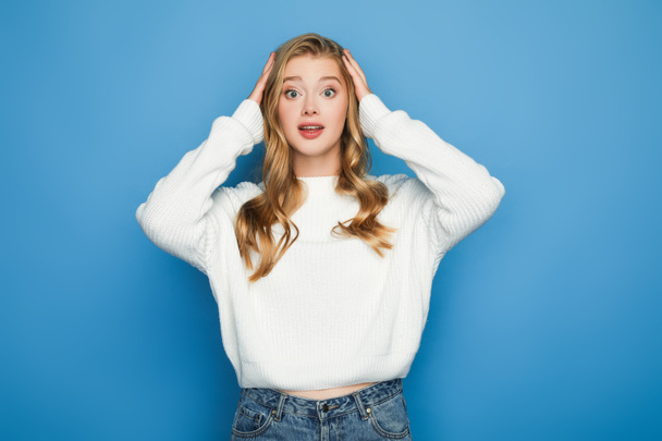 shocked blonde beautiful woman in sweater holding head isolated on blue - 写真・画像
