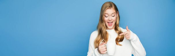 happy blonde beautiful woman in sweater pointing at mug isolated on blue background, banner - Foto, Bild