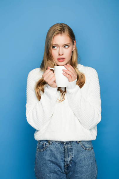 worried blonde beautiful woman in sweater with mug isolated on blue background - Φωτογραφία, εικόνα