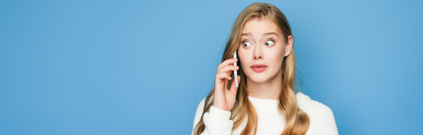 surprised blonde beautiful woman in sweater talking on smartphone isolated on blue background, banner - Fotografie, Obrázek