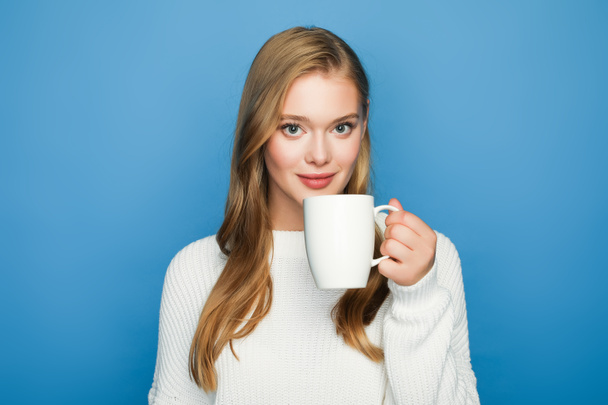 smiling blonde beautiful woman in sweater with mug isolated on blue background - Fotó, kép