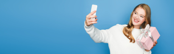 blonde beautiful woman in sweater taking selfie with gift box isolated on blue, banner - Fotoğraf, Görsel
