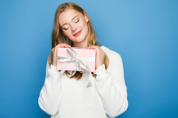 pleased blonde beautiful woman in sweater with gift box isolated on blue - Foto, afbeelding