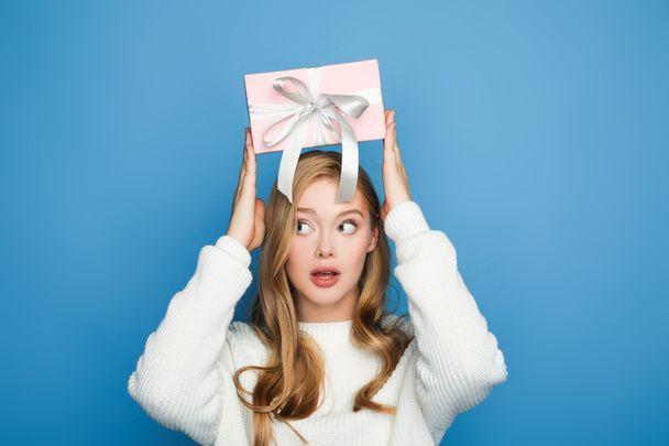 surprised blonde beautiful woman in sweater holding gift box above head isolated on blue - Photo, Image