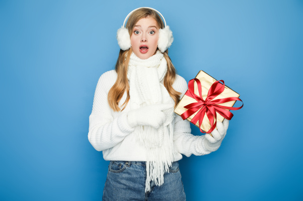 surprised beautiful woman in winter white outfit with gift box isolated on blue - Фото, зображення