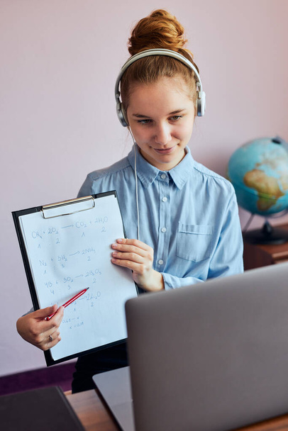 Młoda studentka pokazująca pracę domową, ucząca się online, zdalnie oglądająca lekcje, słuchająca profesora, rozmawiająca z kolegami z klasy na wideo rozmowie z domu podczas kwarantanny. Młoda dziewczyna korzystająca z laptopa, słuchawek, książek, podręczników siedzi przy - Zdjęcie, obraz