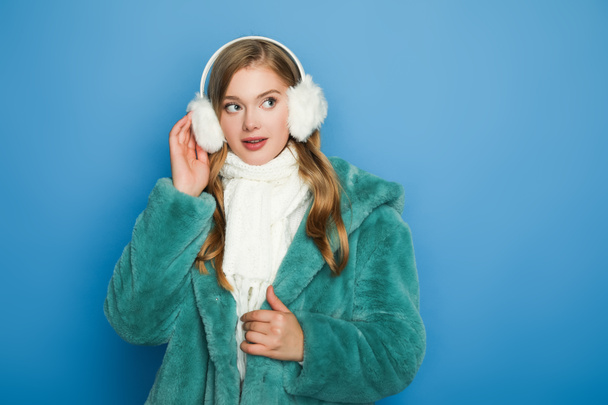 stylish woman in green faux fur coat and ear muffs isolated on blue - Photo, Image