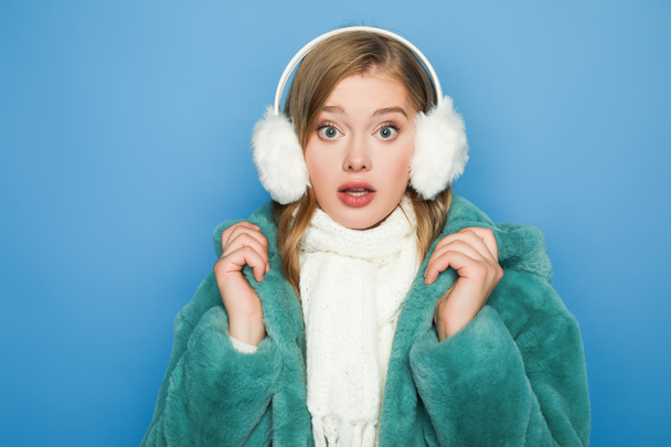 surprised stylish woman in green faux fur coat and ear muffs isolated on blue - Zdjęcie, obraz