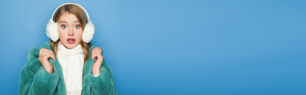 mujer con estilo sorprendido en abrigo de piel sintética verde y orejeras aisladas en azul, pancarta - Foto, Imagen