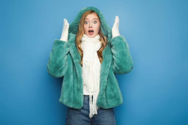 shocked stylish woman in green faux fur coat looking away isolated on blue - Photo, Image