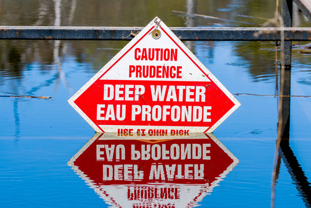 Varování před hlubokou vodou. Část signálu je ponořena. Podpis je v angličtině a francouzštině. Zrcadlení znamení je ve vodě dole. - Fotografie, Obrázek