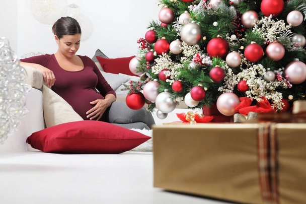 Mutlu noeller, aydınlanmış ve süslenmiş ağacın yanındaki mutlu hamile kadın, altında hediye paketleri, neşeli ve gülümseyerek oturma odasında oturuyor. - Fotoğraf, Görsel