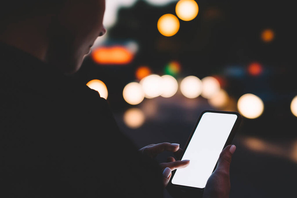 Кадрирование неузнаваемой женщины с помощью мобильного телефона и прикосновение к пустому дисплею с белым экраном, стоя на улице в ночное время - Фото, изображение