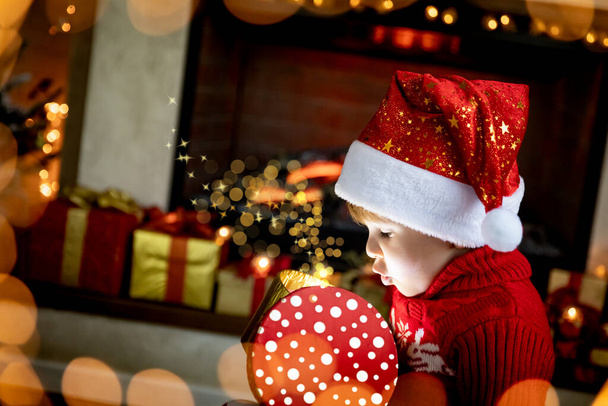 Surprised child opening Christmas gift. Funny kid at home near fireplace. Xmas holiday concept - Fotoğraf, Görsel