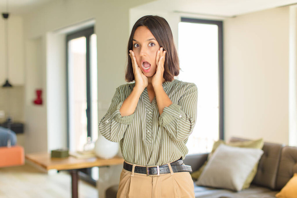 young latin woman feeling shocked and scared, looking terrified with open mouth and hands on cheeks. home concept - 写真・画像