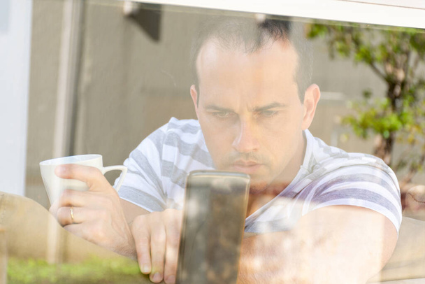 Mann mittleren Alters hinter Fensterglas hält eine Tasse Kaffee in der Hand und benutzt sein Handy. - Foto, Bild