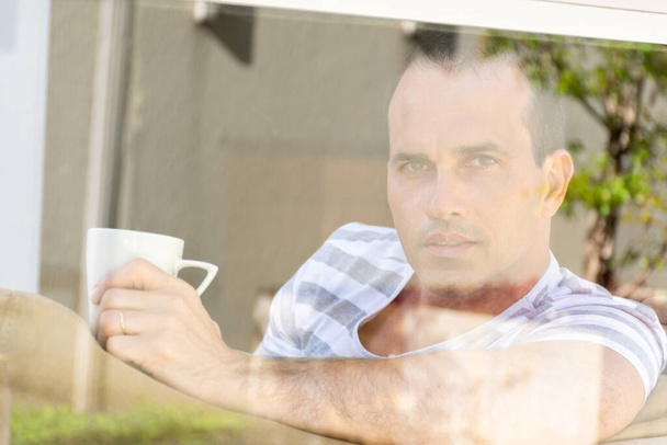 Hombre de mediana edad detrás del cristal de la ventana sosteniendo una taza de café y mirando a la cámara. - Foto, Imagen