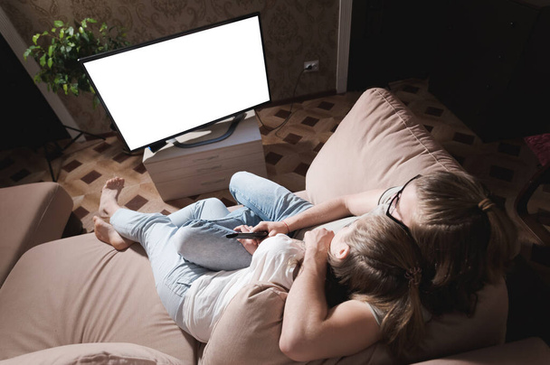 Young family of long haired man and woman are lying on the couch and watching TV. The TV screen is cut out white. Mockup and template for designer layout - Foto, Imagen