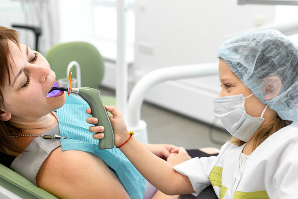 A menina trocou de papel com a dentista. Criança caucasiana segurando equipamentos dentários curadores de luz em suas mãos, usando uma máscara médica, um boné e um manto - Foto, Imagem